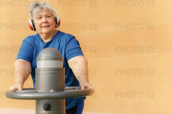 Active obese elderly woman in sportswear working out on a gym machine in a park gym while listening to music on headphones