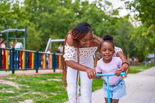 Black African ethnicity mother having fun with her daughter in playground learning to ride a skateboard