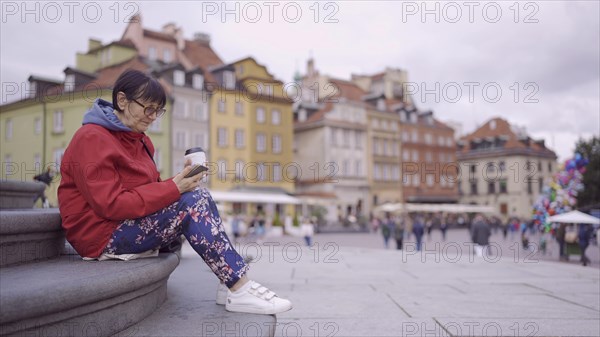 Elderly lady sits on the steps drinking coffee and using a smartphone in the historic center of an old European city. Palace Square