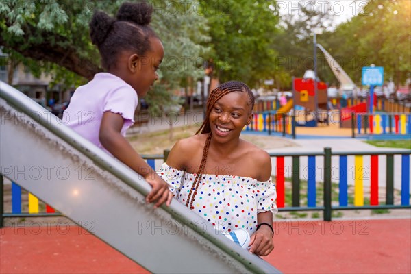 African black ethnicity mother having fun with her daughter in the squeaker of the playground of the city park in the sunset