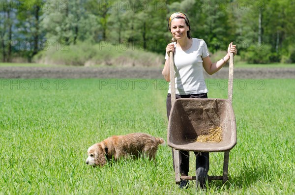 Woman with a Wheelbarrow on the Green Field with Grass and a Dog