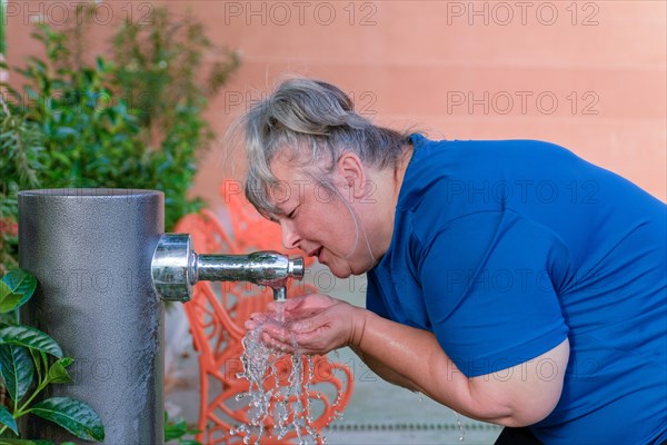 Older white-haired woman in sportswear drinking water from a fountain after workout