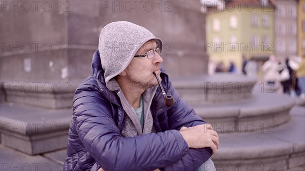 Portrait of adult man with glasses sitting in the hood on square and smoking a tobacco pipe in the Palace Square