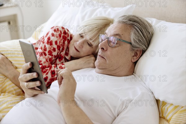 Elderly couple lying together in bed in bedroom looking at smartphone together