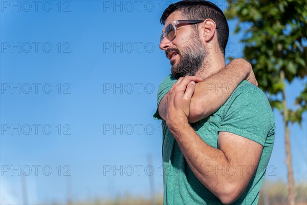 Profile view of an attractive bearded man with sunglasses stretching his shoulders in a field with blue sky in the background