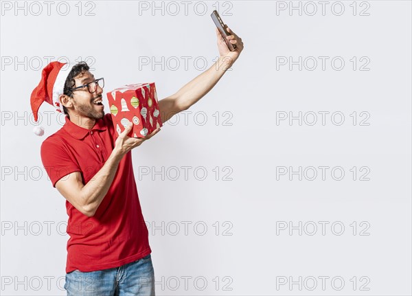 Smiling young man in christmas hat holding a gift and taking a selfie isolated. Christmas handsome man taking a selfie holding a gift isolated. Happy guy taking a christmas selfie isolated