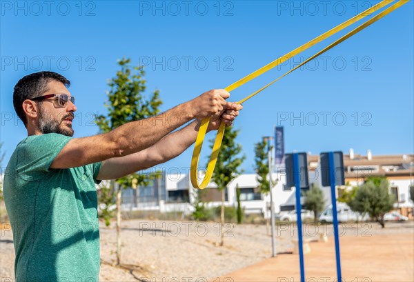 Bearded man with sunglasses seen in profile training his arms with an elastic fitness band in an outdoor gym