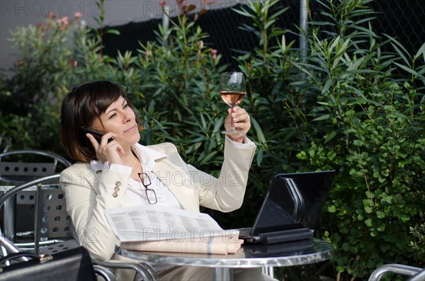 Business Woman Sitting with Her Personal Computer and Financial Newspaper and using Smart Phone on a Table Outdoor