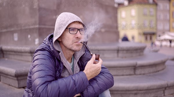 Adult man with glasses sitting on square and smoking a tobacco pipe releasing smoke in the Palace Square