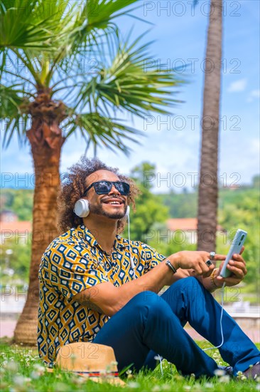 Afro haired man sitting on the grass on summer vacation next to some palm trees by the beach. Traveling and tourism concept