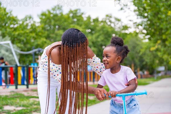 African black ethnicity mother having fun with her daughter in playground walking with the skateboard