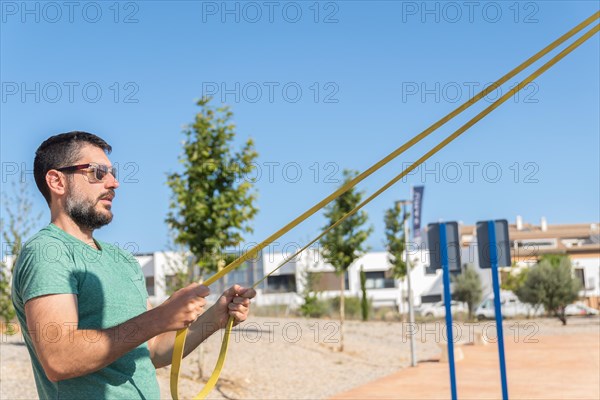 Bearded man with sunglasses seen in profile training his arms with an elastic fitness band in an outdoor gym