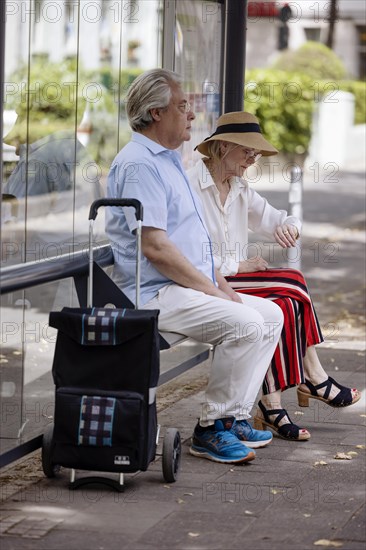 Older couple dressed in summer clothes with a trolley waiting for a bus at a bus stop