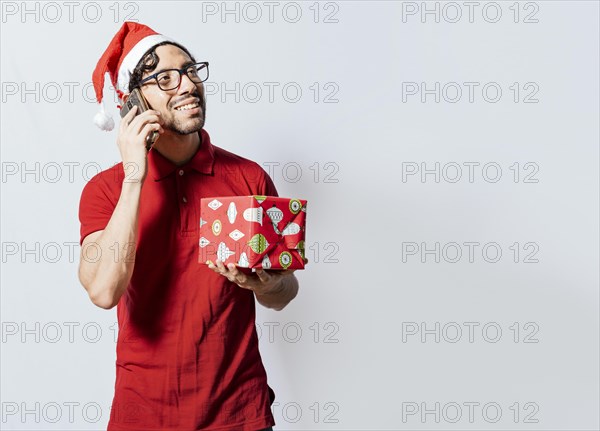 Smiling guy in christmas hat holding christmas gift box and talking on the phone. Christmas man holding gift and calling family on phone. Happy man holding gift and calling family at christmas