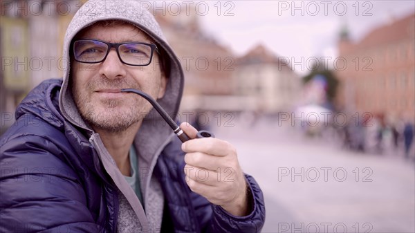 Portrait of adult man with glasses sitting in the hood on square and smoking a tobacco pipe in the Palace Square