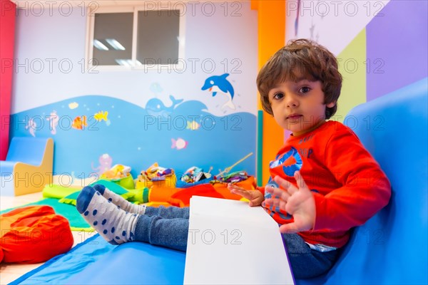 A boy sitting on a blue sofa reading a story book looking at the camera