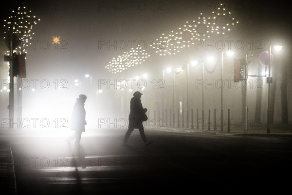 Two people walk along a street decorated with Christmas lights in the fog in Niesky