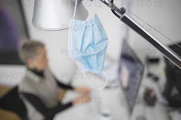 Mask at the workplace. A mask hangs from a lamp above a desk while a woman works on a computer in the background. Berlin