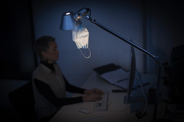 Mask at the workplace. A mask hangs from a lamp above a desk while a woman works on a computer in the background. Berlin