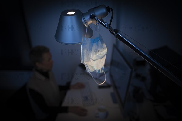 Mask at the workplace. A mask hangs from a lamp above a desk while a woman works on a computer in the background. Berlin