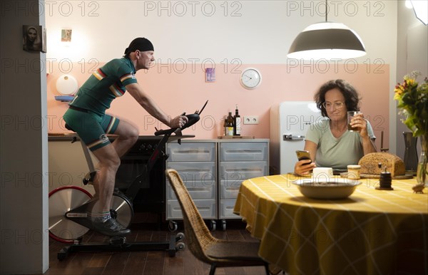 Man on a spinning bike in his kitchen while the woman is sitting at the table