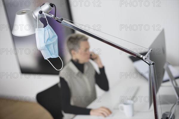 Mask at the workplace. A mask hangs from a lamp above a desk while a woman works on a computer in the background. Berlin