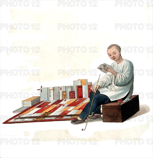 A Chinese bookseller sitting on a box in the street reading in front of his books