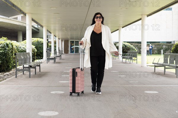 Mature woman in white cover-up leaving terminal and carrying her pink suitcase on wheels. Concept of business travel.Concept of business travel