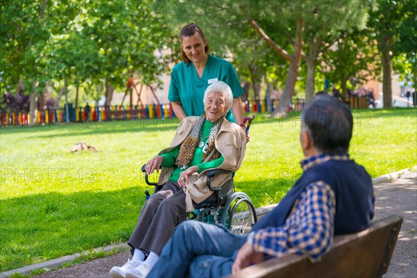 An elderly woman with the nurse on a walk in the garden of a nursing home in a wheelchair in nature and greeting an elderly man