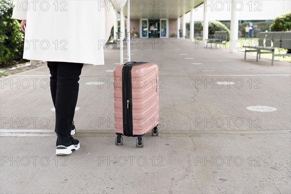 Shot of legs of woman in sneakers and coat walking to main entrance of airport and rolling her wheeled suitcase. Travel concept