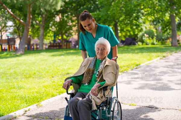 An elderly woman with the nurse on a walk through the garden of a nursing home in a wheelchair next to nature and trees
