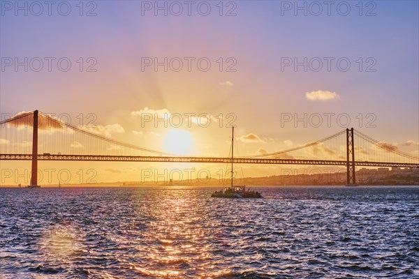 View of 25 de Abril Bridge famous tourist landmark of Lisbon connecting Lisboa and Almada on Setubal Peninsula over Tagus river with tourist yacht silhouette at sunset. Lisbon