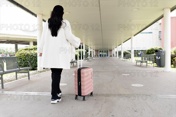 Back shot of woman in sneakers and coat walking towards main entrance of airport and rolling her wheeled suitcase. Travel concept