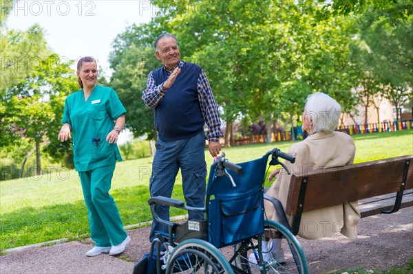 An elderly man with the nurse on a walk through the garden of a nursing home greeting an elderly woman in a wheelchair
