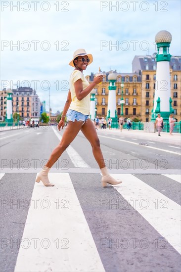 Portrait of black ethnic woman drinking an orange juice enjoying summer on vacation crossing the street at the zebra crossing