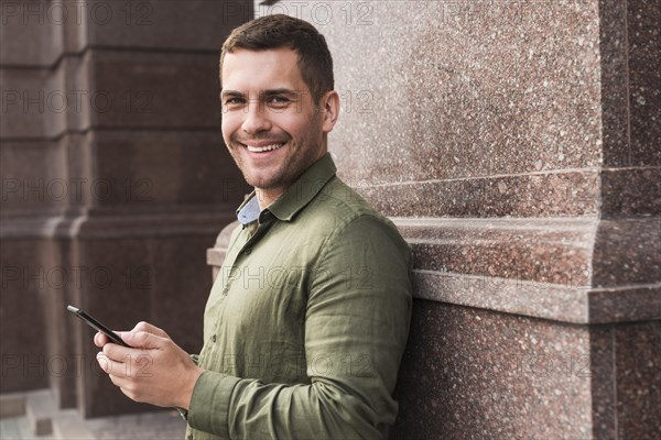 Smiling man leaning wall holding cellphone looking camera