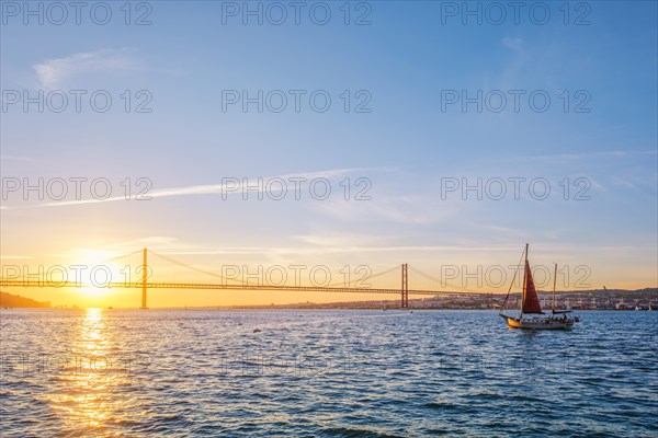 View of 25 de Abril Bridge famous tourist landmark of Lisbon connecting Lisboa and Almada over Tagus river with tourist yacht silhouette at sunset. Lisbon