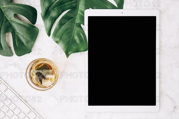 Brewing hot beverage with tea bag glass monstera leaves keyboard digital tablet white desk