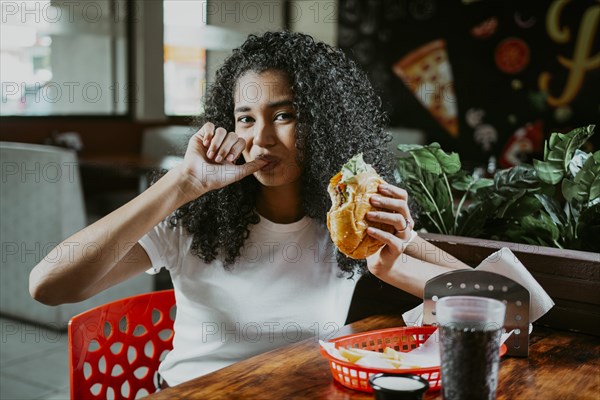 Portrait of an afro girl enjoying hamburger in a restaurant. Latin woman sucking her fingers holding a hamburger in a restaurant