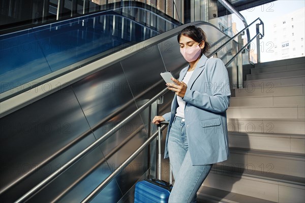 Woman with medical mask luggage using smartphone while climbing down stairs during pandemic