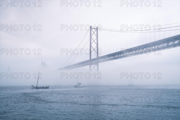 View of 25 de Abril Bridge famous tourist landmark of Lisbon connecting Lisboa and Almada in heavy fog mist wtih yacht boats passing under. Lisbon