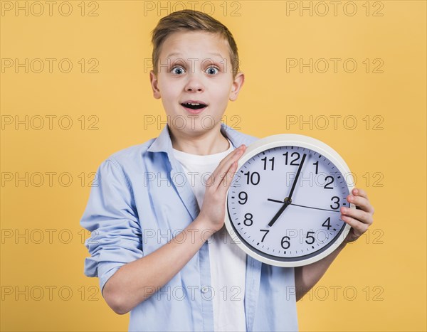 Surprise smiling boy holding white clock hand looking camera against yellow background
