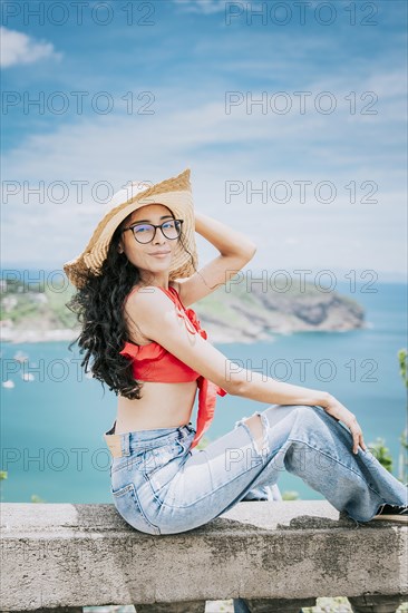 Beautiful tourist girl in a viewpoint with a beach in the background. Happy tourist sitting at a viewpoint looking at the camera with bay in the background