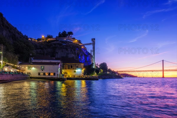 View of 25 de Abril Bridge famous tourist landmark of Lisbon over Tagus river and Boca do Vento Elevator and seaside restaurants with people in the evening. Lisbon