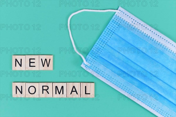 Flat lay with blue disposable protective face mask and wooden letters forming words 'new normal' on teal background