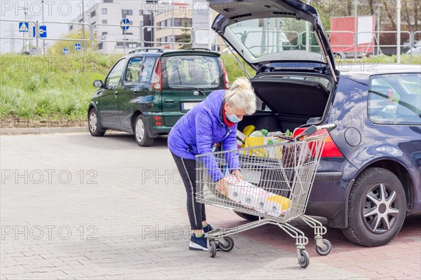 A mature good looking woman wearing protective mask loads toilet paper into a car in the parking lot. Everyday life during coronavirus pandemic. Shopping concept