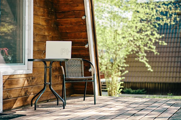 Laptop on the coffee table near whicker chair on terrace of log cabin. Concept of remote work and freelance