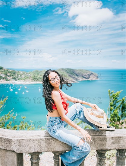 Lifestyle of a tourist girl with a hat in a viewpoint with a beach in the background. Happy female tourist sitting at a viewpoint with beach in the background