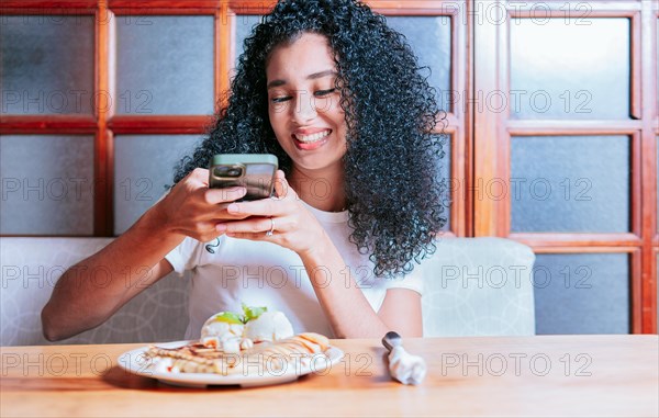 Beautiful girl sitting photographing a chocolate crepe and ice cream. Smiling woman taking a photo of a chocolate crepe on table