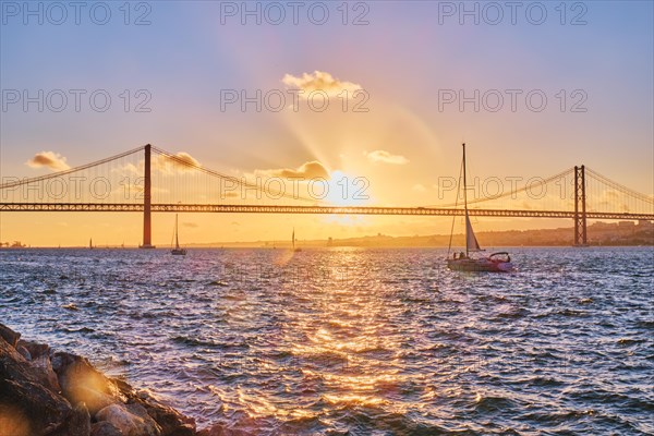 View of 25 de Abril Bridge famous tourist landmark of Lisbon connecting Lisboa and Almada on Setubal Peninsula over Tagus river with tourist yacht silhouette at sunset. Lisbon